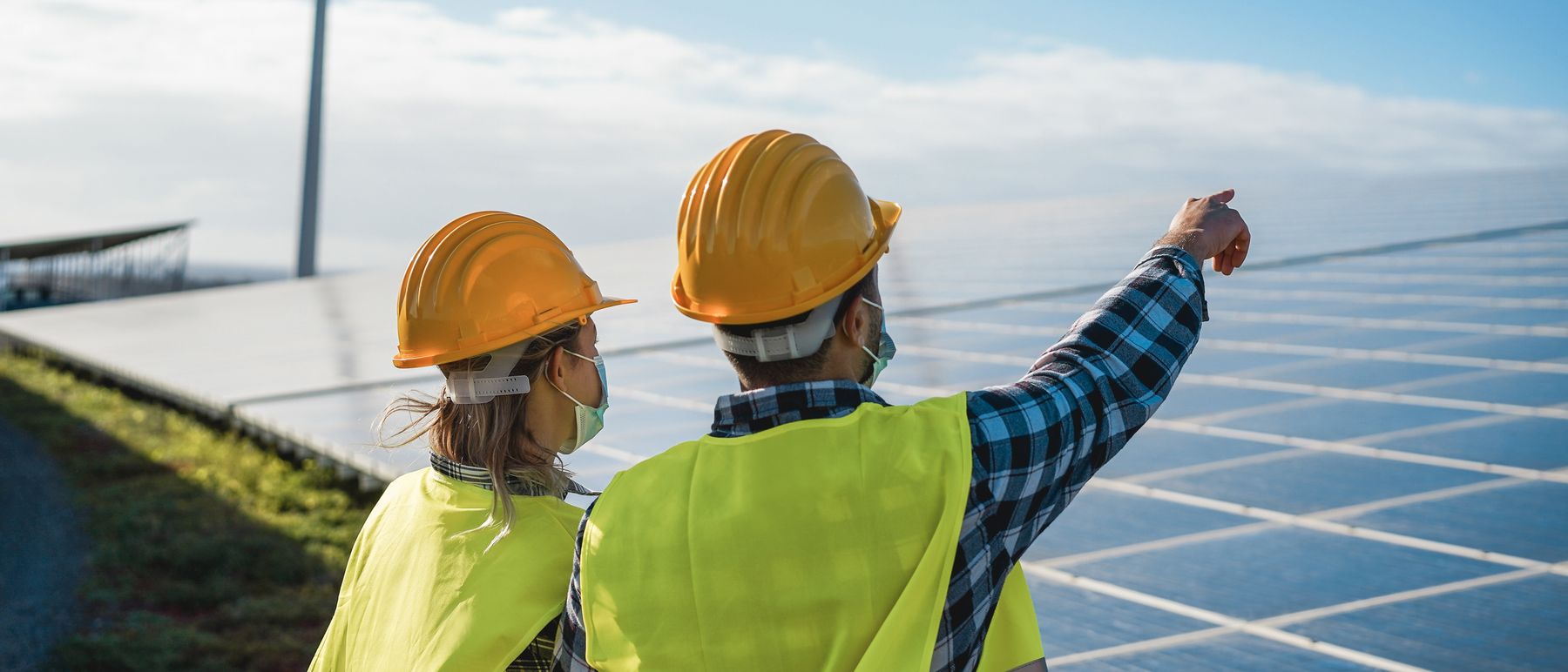 Dvoje inženjera s kacigama pregledava solarnu elektranu, vjetroturbina u pozadini